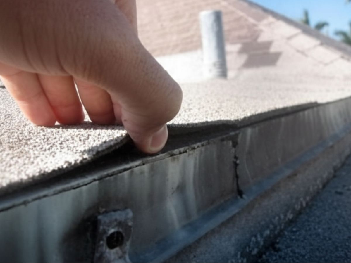 Hand lifting loose asphalt shingle at parapet flashing, showing improper sealing and potential water intrusion point