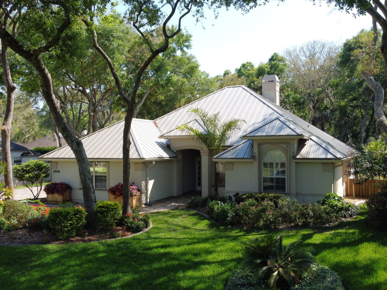 Front exterior view of a single-story home with a light-colored standing seam metal roof, landscaped yard, and mature trees.