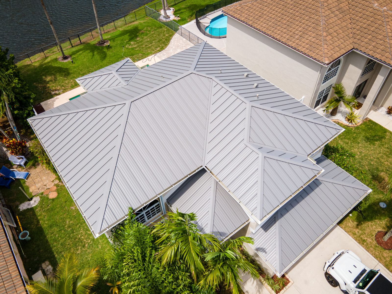 Aerial view of a residential home with a gray standing seam metal roof, multiple roof planes, and surrounding palm trees near a waterfront property