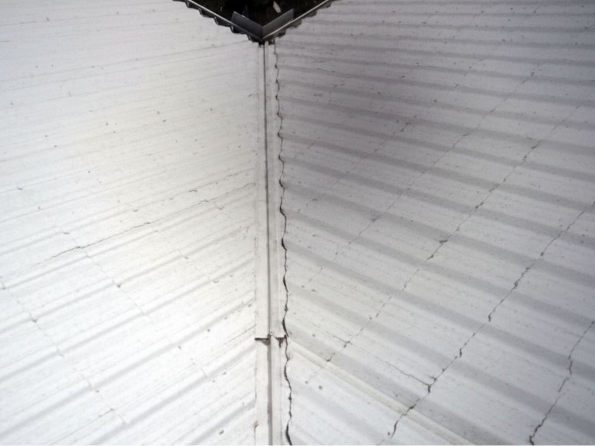 Cracked roof seam at the intersection of two corrugated metal panels, showing separation along the vertical joint.