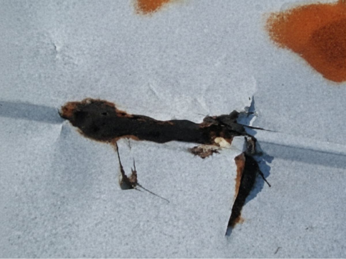Rust-through damage on a metal roof panel with visible corrosion, holes, and deteriorated coating.