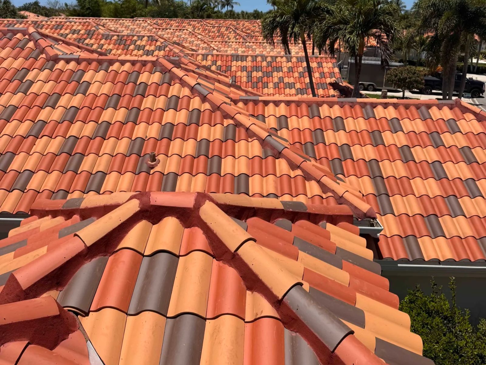 Angled rooftop view of a multicolored clay tile roof with terracotta, tan, and charcoal tiles, featuring hip and ridge lines, mortar detailing, and palm trees visible in the surrounding residential area.