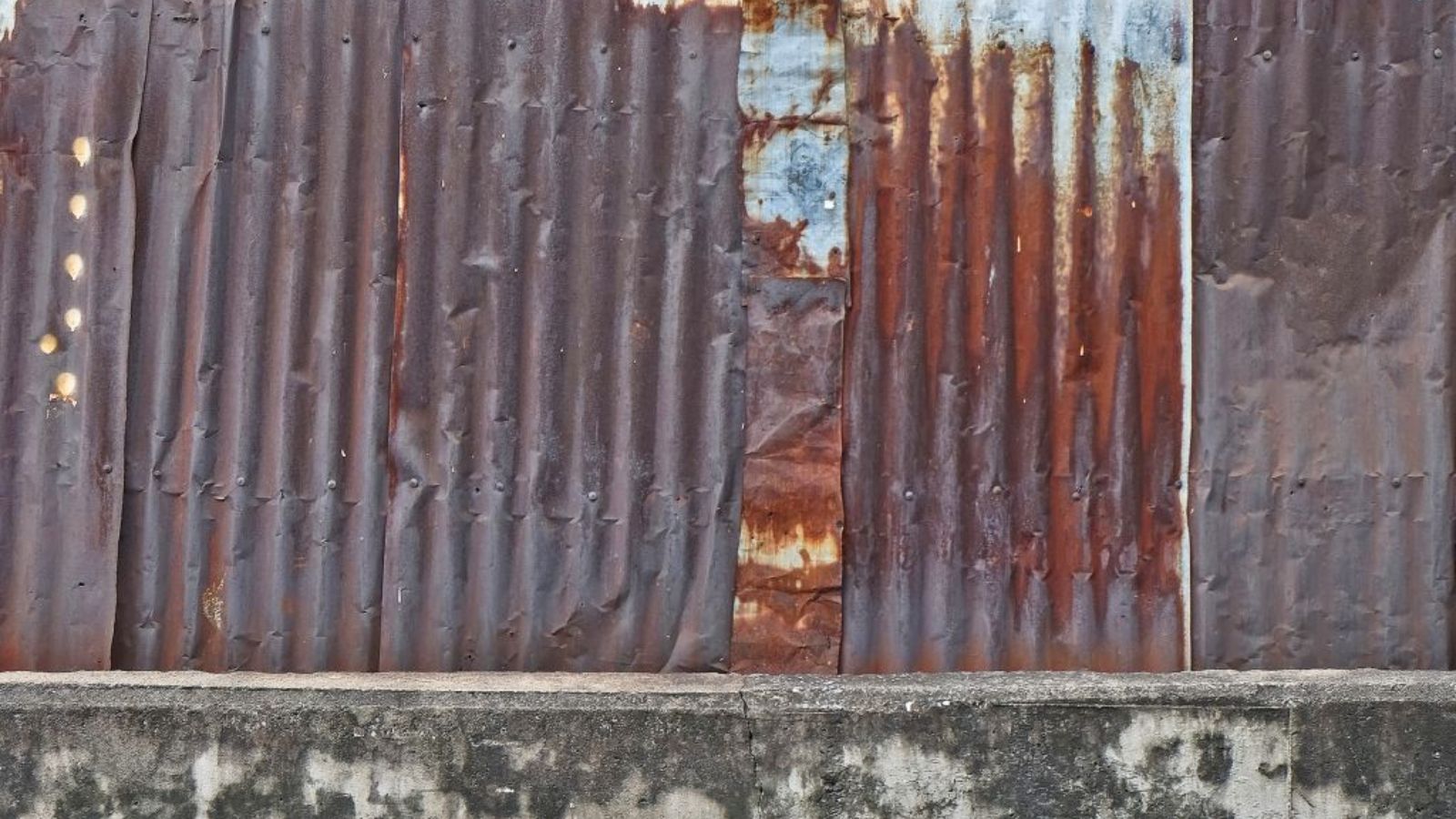 Rusted corrugated metal wall panels with heavy corrosion, dents, and patchwork sections above a concrete base