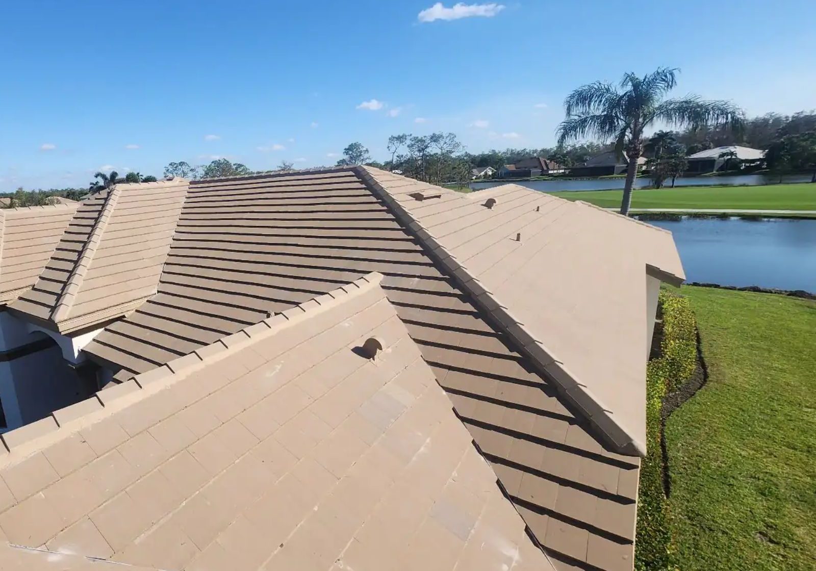 An elevated view of a tan concrete tile roof on a residential home, showing multiple roof planes with clean ridgelines and uniform tile spacing, set against a landscaped backyard with a pond, palm trees, and a clear blue sky.